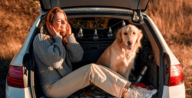 Köpeğiyle sıcak bir battaniyenin üzerinde açık bir arabanın bagajında oturmuş kulaklıkla müzik dinleyen mutlu bir kadın. Evcil hayvanla gezen kadın.