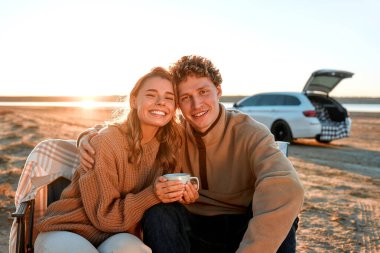 Birlikte oturup kahve ya da çay içen bir çift. Arka planda bagajı açık bir araba var. Aile seyahati. Seyahat, tatil ve turizm konsepti.