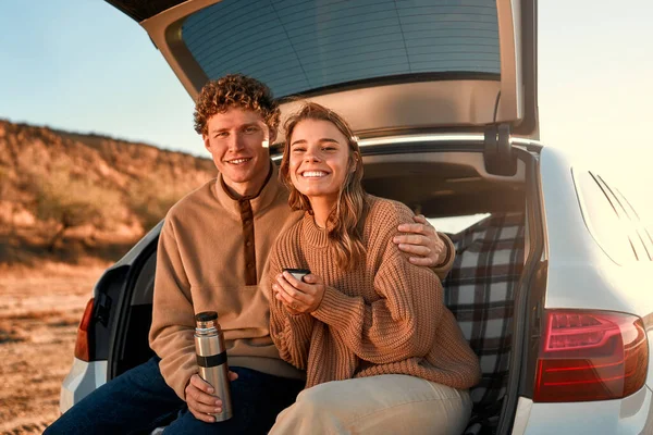 愛する幸せな若いカップルは 暖かい毛布の上の車のオープントランクに座って 週末にサーモスからお茶を飲んでいます 旅行と休暇のコンセプト — ストック写真