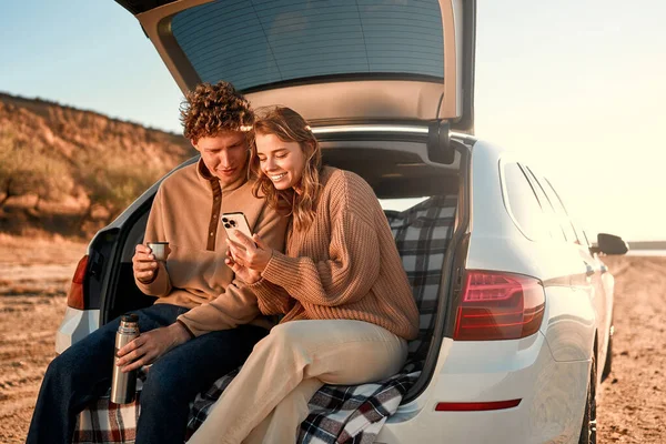 愛する幸せな若いカップルは 暖かい毛布の上の車のオープントランクに座って サーモスからお茶を飲んで 週末にスマートフォンを使用しています 旅行と休暇のコンセプト — ストック写真