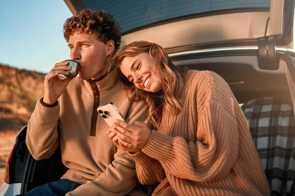 一对恋爱中的快乐的年轻夫妇在温暖的毛毯上坐在汽车的敞篷后备箱里 喝着热水瓶里的茶 周末还使用智能手机 旅行和休假概念 — 图库照片