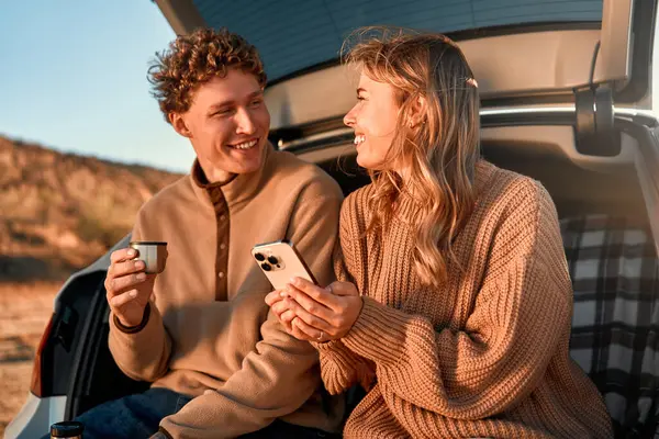 愛する幸せな若いカップルは 暖かい毛布の上の車のオープントランクに座って サーモスからお茶を飲んで 週末にスマートフォンを使用しています 旅行と休暇のコンセプト — ストック写真