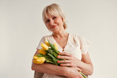 Anneler ve kadınlar günün kutlu olsun. Beyaz bir arka planda izole lale demeti olan bir elbise giymiş, çekici, olgun bir kadın. Annenin sevgisi ve bakımı.