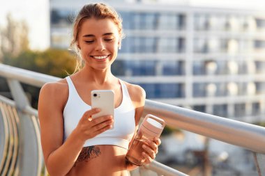 Elinde bir şişe su olan bir kadın köprüde açık havada koştuktan sonra rahatlarken akıllı telefon kullanıyor. Sabah egzersizleri ve koşu. Spor ve eğlence, aktif yaşam tarzı.