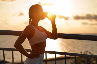 Denizin arka planına karşı bir köprüde koştuktan sonra dinlenirken su içen bir kadın. Sabah egzersizleri ve koşu. Spor ve eğlence, aktif yaşam tarzı.