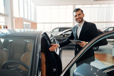 Takım elbiseli ve tabletli bir satıcı bir araba galerisinde durur ve müşterilere koltukları göstererek arabayı gösterir. Almak, kiralamak ve bir arabayı sigortalamak..