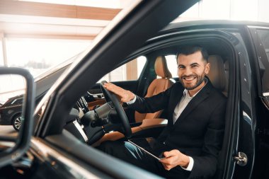 Tabletli takım elbiseli bir satıcı araba galerisinde durur ve arabayı alıcılara gösterir, direksiyonu gösterir. Almak, kiralamak ve bir arabayı sigortalamak..