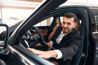 Takım elbiseli bir satıcı, elinde tabletle galeride duruyor ve alıcılara araba gösteriyor. Almak, kiralamak ve bir arabayı sigortalamak..