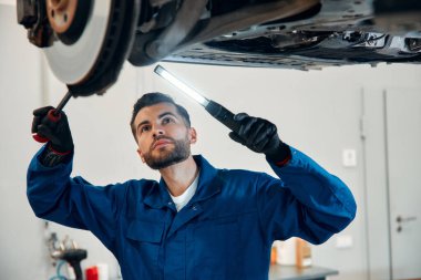 Yoğunlaştırılmış araba servisi çalışanının otomobil kontrol ekipmanlarına bakın. Üniformalı ve eldivenli erkek tamirci arabanın altında duruyor, lambayı tutuyor ve kırılma noktası arıyor..