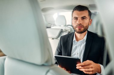 Siyah takım elbiseli, beyaz derili lüks bir arabada oturan ve tablet kullanan bir erkek işadamı..