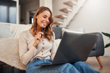 Evdeki oturma odasındaki kanepede otururken dizüstü bilgisayarda çalışan genç bir kadın. Uzak çalışma ve serbest çalışma.