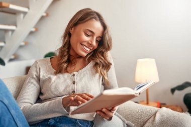 Kanepede kitap okuyan genç bir kadın. Evdeki rahat bir oturma odasında. İzin gününde dinleniyor ve dinleniyor..