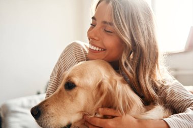 Evdeki rahat oturma odasında köpeği olan mutlu genç bir kadın hafta sonu rahatlıyor ve evcil hayvanıyla sarılıp oynuyor..