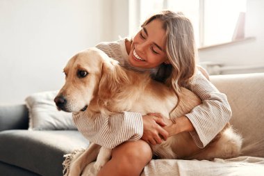 Evdeki rahat oturma odasında köpeği olan mutlu genç bir kadın hafta sonu rahatlıyor ve evcil hayvanıyla sarılıp oynuyor..