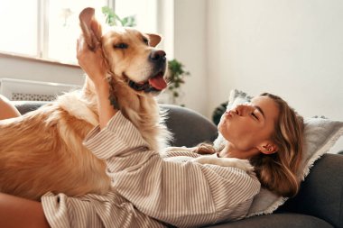 Evdeki rahat oturma odasında köpeği olan mutlu genç bir kadın hafta sonu rahatlıyor ve evcil hayvanıyla sarılıp oynuyor..