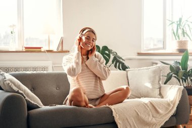 Evde oturma odasındaki kanepenin yanında otururken müzik ya da kulaklık dersi alan genç bir kadın..