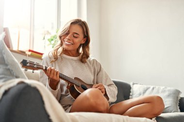 Mutlu genç bir kadın, hafta sonları evdeki rahat oturma odasında oturup dinlenirken gitar çalıyor..
