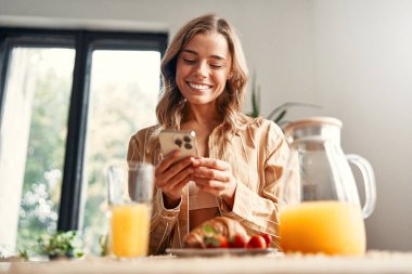 Mutfaktaki masada duran genç mutlu kadın akıllı telefon kullanıyor. Tabakta çilekli kruvasan ve bir bardak meyve suyu var. Sıcak bir mutfakta kahvaltı yapan bir kadın..