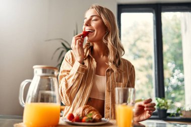 Mutfaktaki masada oturan, çilekli kruvasan yiyen ve meyve suyu içen genç ve mutlu bir kadın. Sıcak bir mutfakta kahvaltı yapan bir kadın..