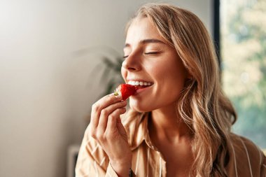 Mutfakta çilek yiyen ve gülümseyen genç bir kadın. Sıcak bir mutfakta kahvaltı yapan bir kadın..