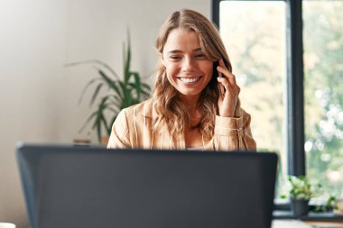 Mutfaktaki masada oturan, dizüstü bilgisayarda çalışan ve akıllı telefon kullanan genç ve mutlu bir kadın. İş kadını, evden uzakta çalışıyor..