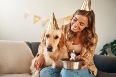 Genç, mutlu bir kadın elinde doğum günü pastası, evcil köpeğinin doğum gününü kutluyor, evcil hayvanıyla kucaklaşıyor ve onunla eğleniyor..