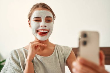 Bir kadın sabah kilden bir maske taktı ve evdeki yatak odasındaki akıllı telefona selfie çekti. Cilt bakımı ve kaplıca tedavisi.