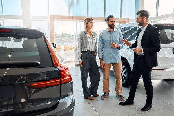 Продавец в костюме с планшетом консультирует пару клиентов в автосалоне. Покупка, аренда и страхование автомобиля.