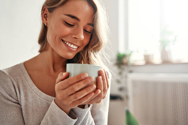 Молодая женщина с чашкой горячего кофе или чая сидит на диване в уютной гостиной дома расслабляясь и раскручиваясь в выходной день.