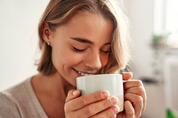 Молодая женщина с чашкой горячего кофе или чая сидит на диване в уютной гостиной дома расслабляясь и раскручиваясь в выходной день.