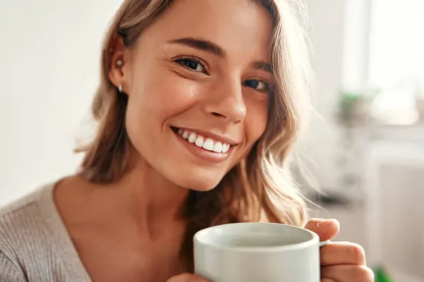 Young woman with a cup of hot coffee or tea sitting on a sofa in a cozy living room at home relaxing and unwinding on a weekend day.