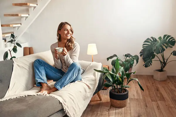 Young woman with a cup of hot coffee or tea sitting on a sofa in a cozy living room at home relaxing and unwinding on a weekend day.