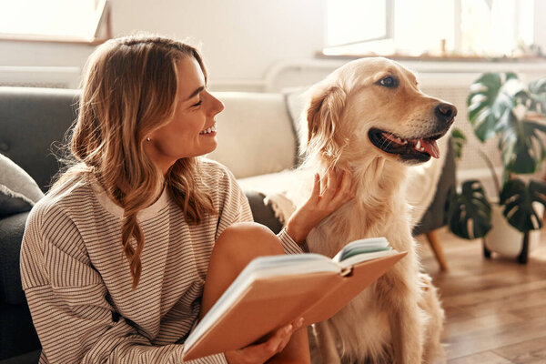 Happy young woman with a dog on the sofa in a cozy living room at home relaxing and resting on a day off, hugging and playing with her pet and reading a book.