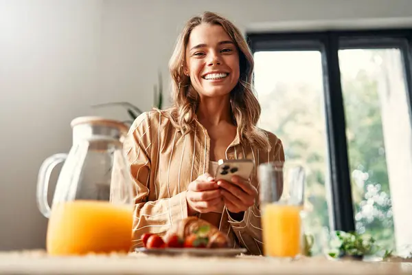Young happy woman standing at the table in the kitchen, using a smartphone, there are croissants with strawberries in a plate, and a glass of juice. Woman having breakfast in a cozy kitchen.