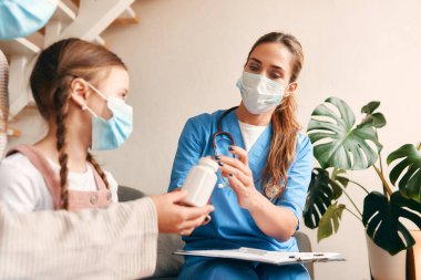 Evde, sağlık maskesi takmış bir aile doktoru oturma odasındaki kanepede otururken bir çocuğa ilaç veriyor. Hasta muayenesi, sağlık ve ilaç.