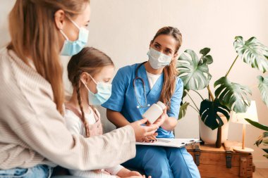 Evde, sağlık maskesi takmış bir aile doktoru oturma odasındaki kanepede otururken bir çocuğa ilaç veriyor. Hasta muayenesi, sağlık ve ilaç.