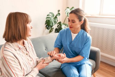 Mutlu genç bayan hemşire, evde bakım ziyaretinde gülümseyen olgun kadınlara tıbbi destek sağlıyor, bayan bakıcı doktor empati gösteriyor emekli hastayı evdeki hastanedeki kanepede oturmaya teşvik ediyor.