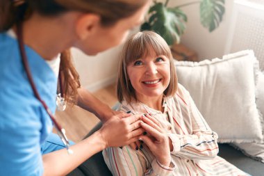 Mutlu genç bayan hemşire, evde bakım ziyaretinde gülümseyen olgun kadınlara tıbbi destek sağlıyor, bayan bakıcı doktor empati gösteriyor emekli hastayı evdeki hastanedeki kanepede oturmaya teşvik ediyor.