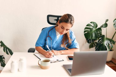 Steteskoplu bir doktor elinde steteskopla dizüstü bilgisayarla bir masada oturup not alıyor ve hastanedeki bir doktorun ofisinde kahve içiyor. Sağlık ve ilaç, çevrimiçi danışma.