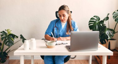 Steteskoplu bir doktor elinde steteskopla dizüstü bilgisayarla bir masada oturup not alıyor ve hastanedeki bir doktorun ofisinde kahve içiyor. Sağlık ve ilaç, çevrimiçi danışma.