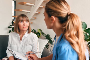 Evde steteskoplu bir aile doktoru oturma odasındaki kanepede oturan yaşlı bir kadını muayene ediyor. Hasta muayenesi, sağlık ve ilaç.