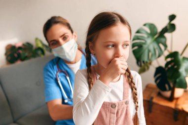 Evde, sağlık maskesi takmış bir aile doktoru oturma odasında bir kanepede otururken bir çocuğun ciğerlerini dinliyor. Hasta muayenesi, sağlık ve ilaç.