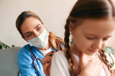 Evde, sağlık maskesi takmış bir aile doktoru oturma odasında bir kanepede otururken bir çocuğun ciğerlerini dinliyor. Hasta muayenesi, sağlık ve ilaç.