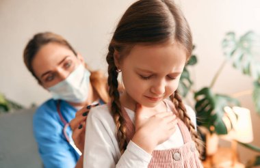 Evde, sağlık maskesi takmış bir aile doktoru oturma odasında bir kanepede otururken bir çocuğun ciğerlerini dinliyor. Hasta muayenesi, sağlık ve ilaç.