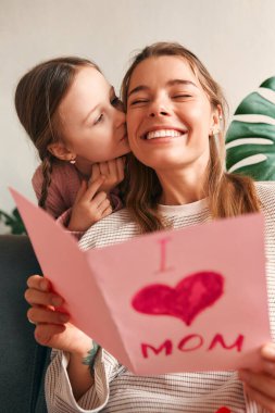 Çocuk kız, oturma odasında kanepede otururken ona bir kart vererek annesini tebrik ediyor. Anneler günü ve kadınlar günü konsepti.