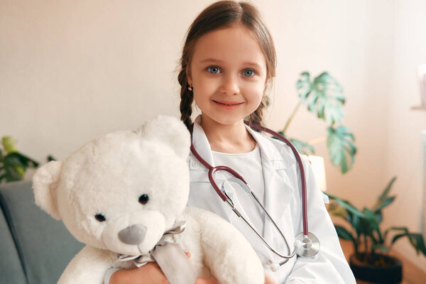 Child girl in a medical gown with a stethoscope treating a teddy bear sitting on the sofa in the living room.