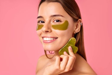 Gözlerinin altında nemlendirici bantları olan güzel bir kadın pembe arka planda Gua sha kazıyıcıyla masaj yapıyor. Yüz bakımı, kozmetoloji, güzellik konsepti..