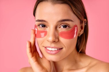 Gözlerinin altında nemlendirici bantları olan güzel bir kadın pembe arka planda izole edilmiş. Yüz bakımı, kozmetoloji, güzellik konsepti..