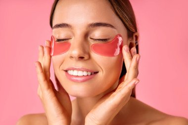 Gözlerinin altında nemlendirici bantları olan güzel bir kadın pembe arka planda izole edilmiş. Yüz bakımı, kozmetoloji, güzellik konsepti..