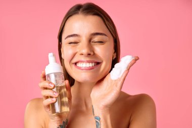 Güzel bir kadın pembe arka planda köpükle yüzünü temizliyor. Yüz bakımı, kozmetoloji, güzellik konsepti..